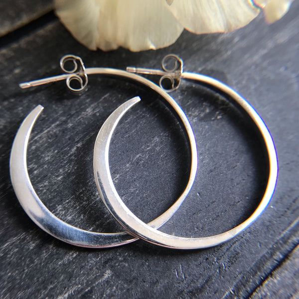 Large sterling silver hoop earrings on a black wood background next to a flower.
